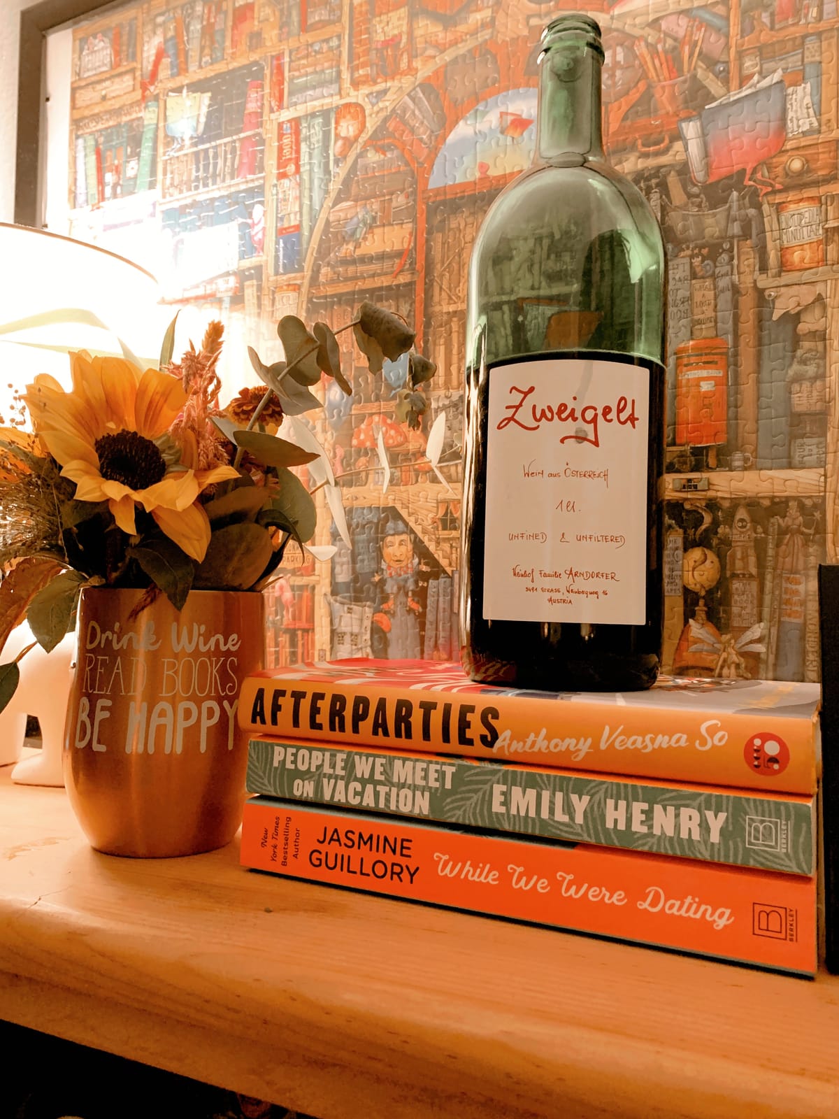 photo of a bookshelf with a wine glass of flowers, a wine bottle, a stack of books, a framed puzzle behind it